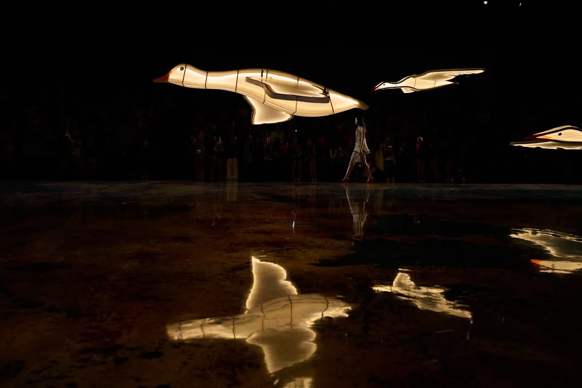 A glowing bird reflected in the dark, water-like runway as a model walks in the distance