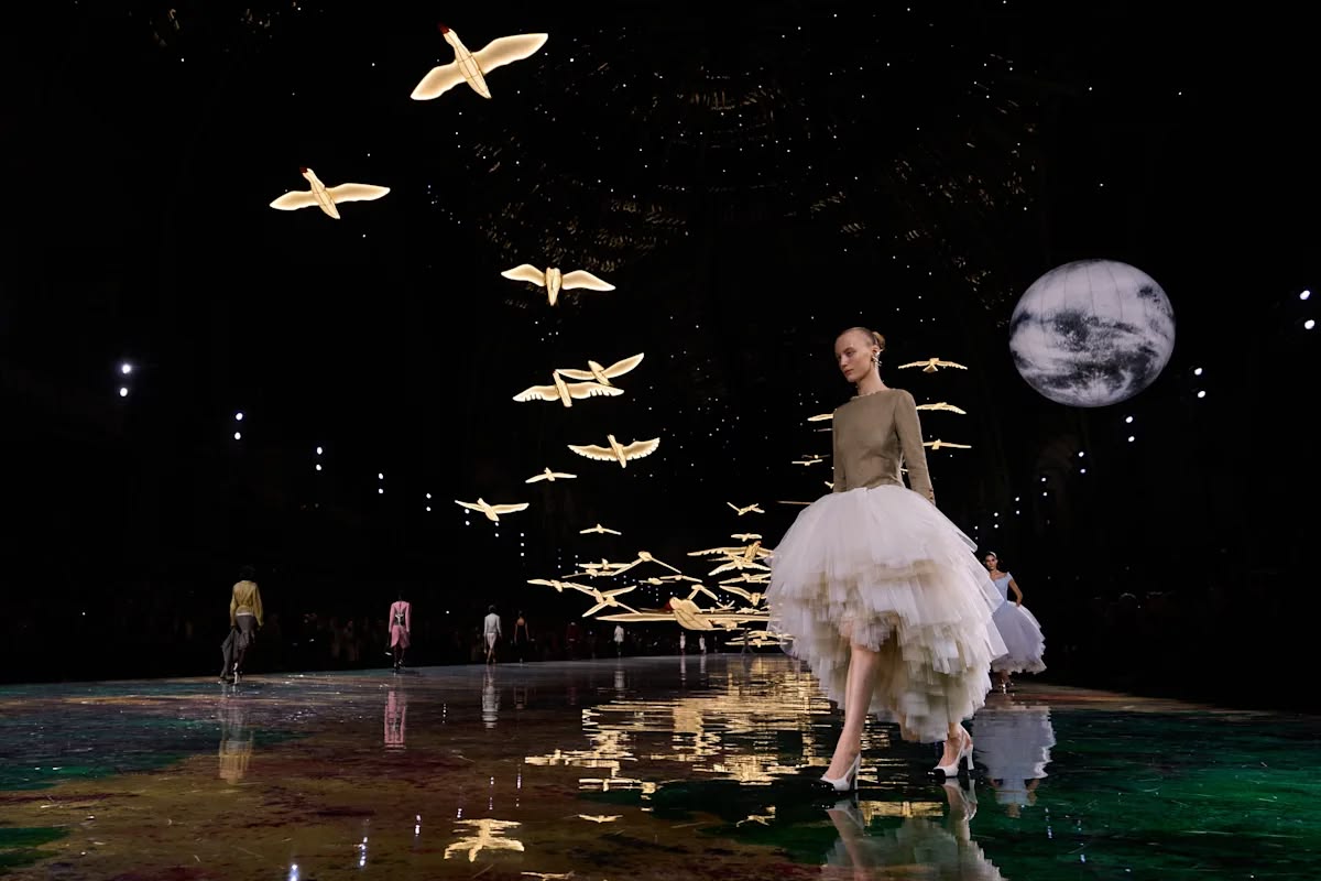 A model in a fitted knit top and voluminous ivory tulle skirt walks beneath the illuminated birds and moon